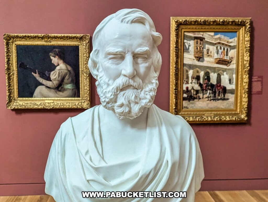 A marble bust of a bearded man on display at the Palmer Museum of Art in Centre County, PA. The sculpture is finely detailed, capturing the subject's thoughtful expression and intricate beard. Behind the bust, two framed paintings are visible: one depicting a woman reading and the other illustrating a lively street scene with horses and people. The artworks are set against a rich, dark red wall, creating a striking contrast with the white marble of the bust. The exhibit highlights the diversity of the museum's collection.