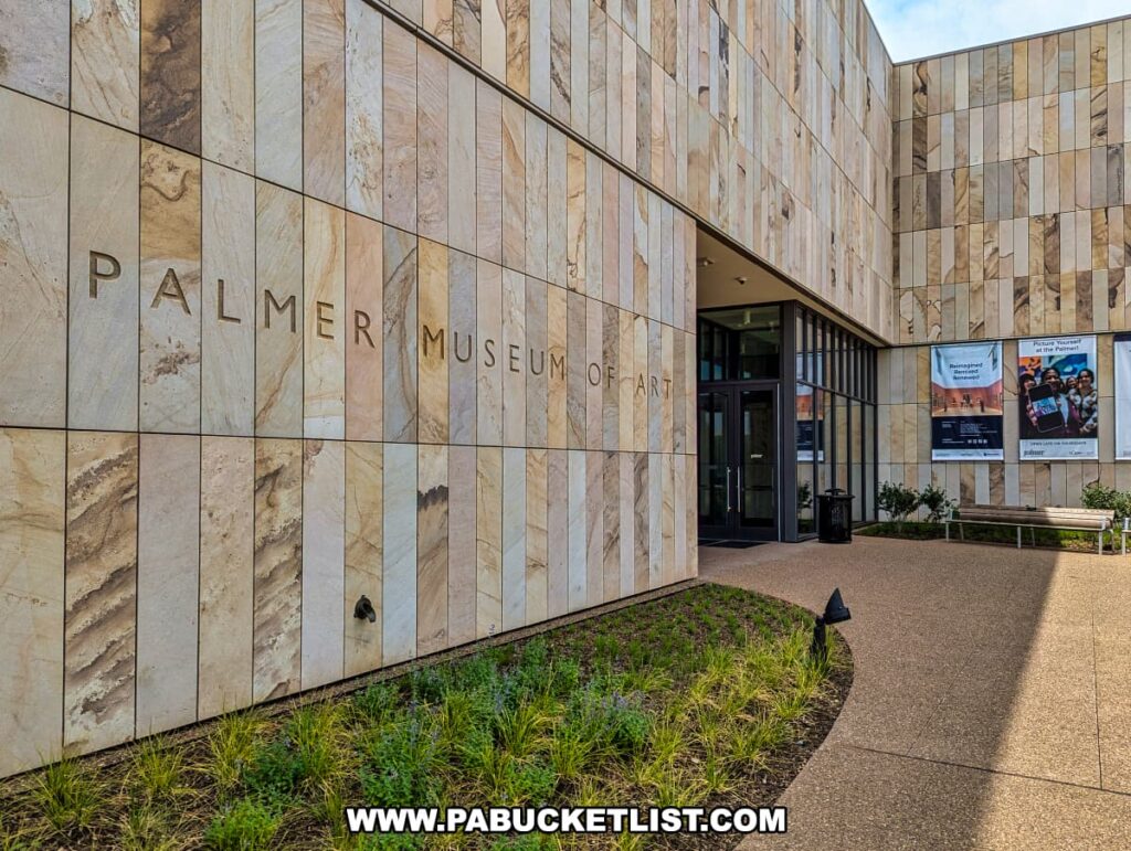 The entrance to the Palmer Museum of Art in Centre County, PA, featuring a modern architectural design with a facade made of light-colored stone tiles. The museum's name is prominently displayed in large, bold letters on the wall. To the right of the entrance, three posters advertise current exhibitions and events. A pathway lined with greenery leads to the glass doors of the museum, inviting visitors inside. A bench is placed near the entrance, providing a spot for rest and contemplation. The overall setting is clean and welcoming, reflecting the museum's significance in the region.