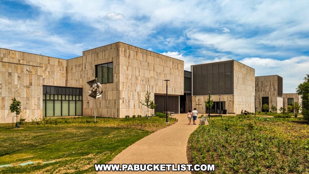An exterior view of the Palmer Museum of Art in Centre County, PA, showcasing its modern architectural design with light-colored stone walls. The building features large windows and geometric shapes, creating a contemporary aesthetic. A pathway leads to the entrance, where two visitors are walking toward the museum. The landscaped grounds surrounding the museum include young trees and well-maintained greenery. A metal sculpture is installed near the entrance, adding an artistic element to the outdoor space. The sky above is partly cloudy, providing a bright and inviting atmosphere.