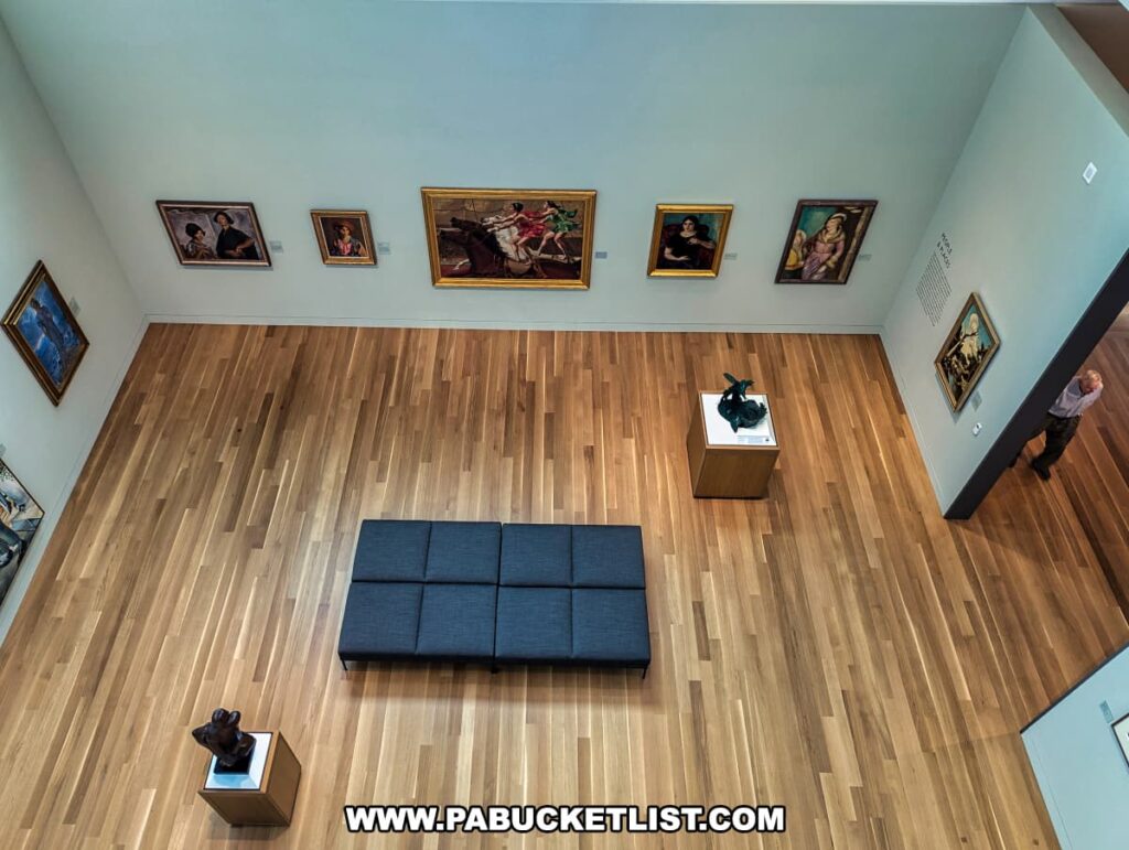 An overhead view of an art gallery at the Palmer Museum of Art in Centre County, PA, showcasing a variety of paintings and sculptures. The gallery features a spacious layout with a wooden floor and light-colored walls. Several framed paintings are displayed on the walls, depicting various subjects including portraits and historical scenes. In the center of the gallery, a gray bench provides seating for visitors. Two sculptures on pedestals are placed strategically within the gallery, adding depth to the exhibition space. A visitor is seen entering the gallery from the right side, enhancing the sense of scale and engagement within the museum.