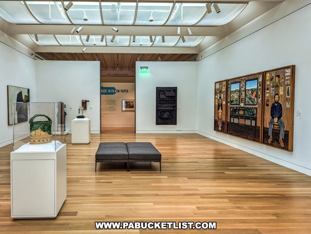 An interior view of one of the galleries at the Palmer Museum of Art in Centre County, PA. The gallery features a variety of artworks, including a large triptych painting on the right wall depicting detailed scenes and portraits. In the foreground, a ceramic basket is displayed in a glass case on a pedestal, and another sculpture is placed on a pedestal nearby. A gray bench provides seating for visitors to admire the art. The room is well-lit with natural light filtering through large, curved skylights in the ceiling, enhancing the bright and airy atmosphere. A sign in the background indicates the entrance to another exhibit titled "Made in PA: On Paper." The wooden floor and white walls create a clean, modern backdrop for the diverse collection of art.