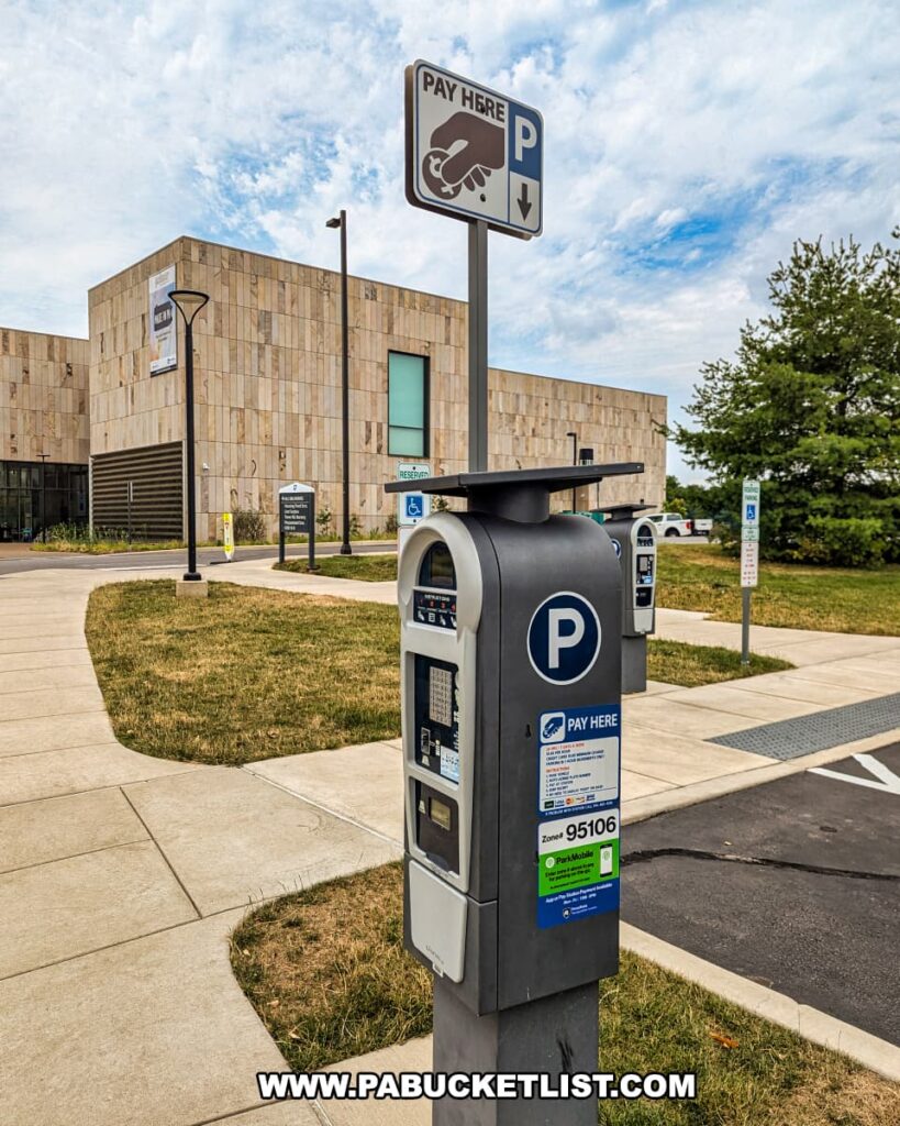 A parking payment station located next to the Palmer Museum of Art in Centre County, PA. The station is equipped with a "Pay Here" sign featuring an icon of a hand inserting coins. The modern, automated machine allows visitors to pay for parking, with instructions and payment options clearly displayed. The background shows the museum's contemporary building with light-colored stone walls and large windows. The area is well-maintained with sidewalks, signage, and nearby greenery, providing a convenient and accessible parking option for museum visitors. The sky is partly cloudy, adding a pleasant atmosphere to the scene.