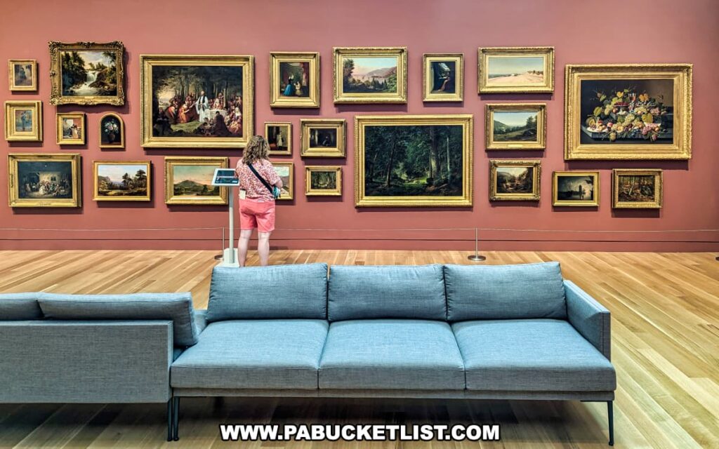 A visitor examines a collection of paintings in a gallery at the Palmer Museum of Art in Centre County, PA. The artworks are arranged in a salon-style display on a red wall, featuring a variety of scenes including landscapes, still lifes, and historical moments, all in ornate gold frames. A gray sofa provides seating in the foreground, offering a comfortable place for visitors to sit and enjoy the art. The wooden floor adds warmth to the space, creating an inviting and reflective atmosphere. The visitor stands in front of an interactive display, enhancing their experience and engagement with the collection.