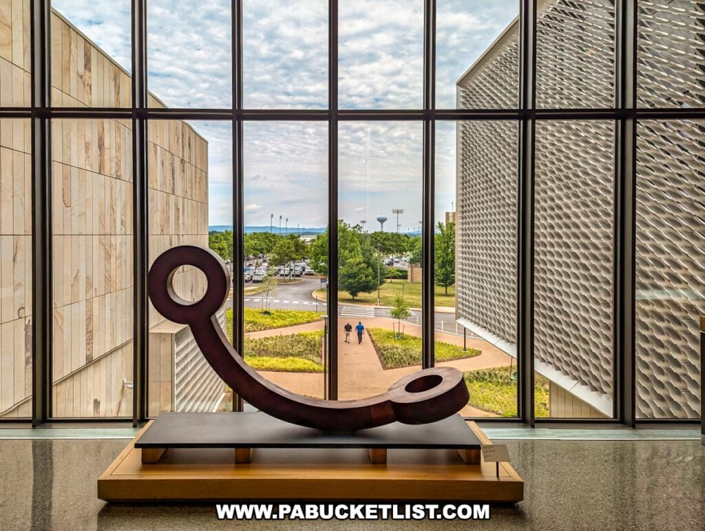 A large sculpture with circular elements is displayed inside the Palmer Museum of Art in Centre County, PA, set against a backdrop of floor-to-ceiling windows. The windows provide a clear view of the exterior landscape, including the museum's entrance pathway, greenery, and parking area. The modern architecture of the museum, with its stone walls and intricate exterior design, frames the outdoor scene. Two people are seen walking along the pathway outside, adding a sense of scale and life to the view. The interior lighting and the natural light from the windows highlight the sculpture's form and the overall spaciousness of the museum's design.