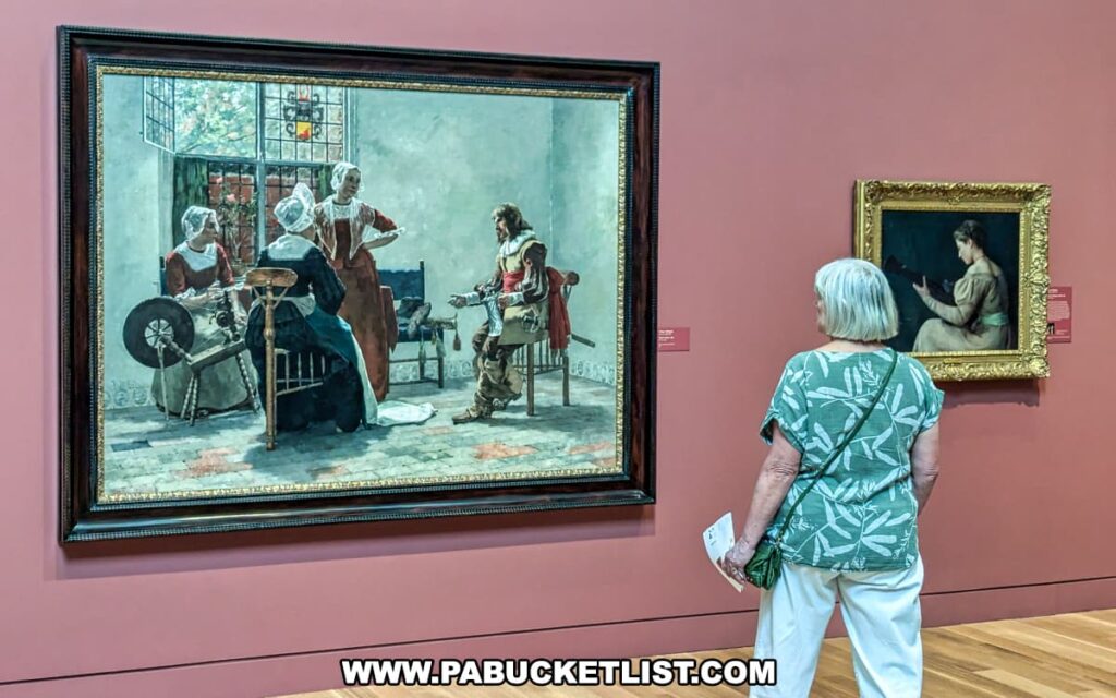 A visitor at the Palmer Museum of Art in Centre County, PA, stands in front of a large painting depicting an interior scene with several figures dressed in historical clothing. The painting, rich in detail, shows women spinning and a man seated, all engaged in a domestic activity. The artwork is framed in an ornate dark frame and set against a red wall. To the right, a smaller painting of a woman reading is displayed in a gold frame. The visitor, wearing a green patterned shirt and white pants, holds a pamphlet and appears to be closely studying the artwork. The wooden floor adds warmth to the gallery space, enhancing the viewing experience.