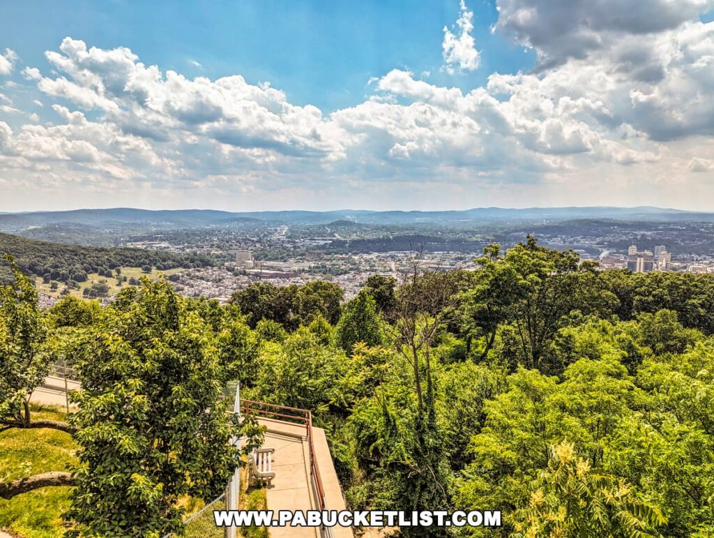 The photo showcases a panoramic view from the Reading Pagoda in Berks County, PA, overlooking the city of Reading and the surrounding landscape. The scene features lush green trees in the foreground, rolling hills, and a vast expanse of the city under a partly cloudy sky, highlighting the beauty and tranquility of the area.