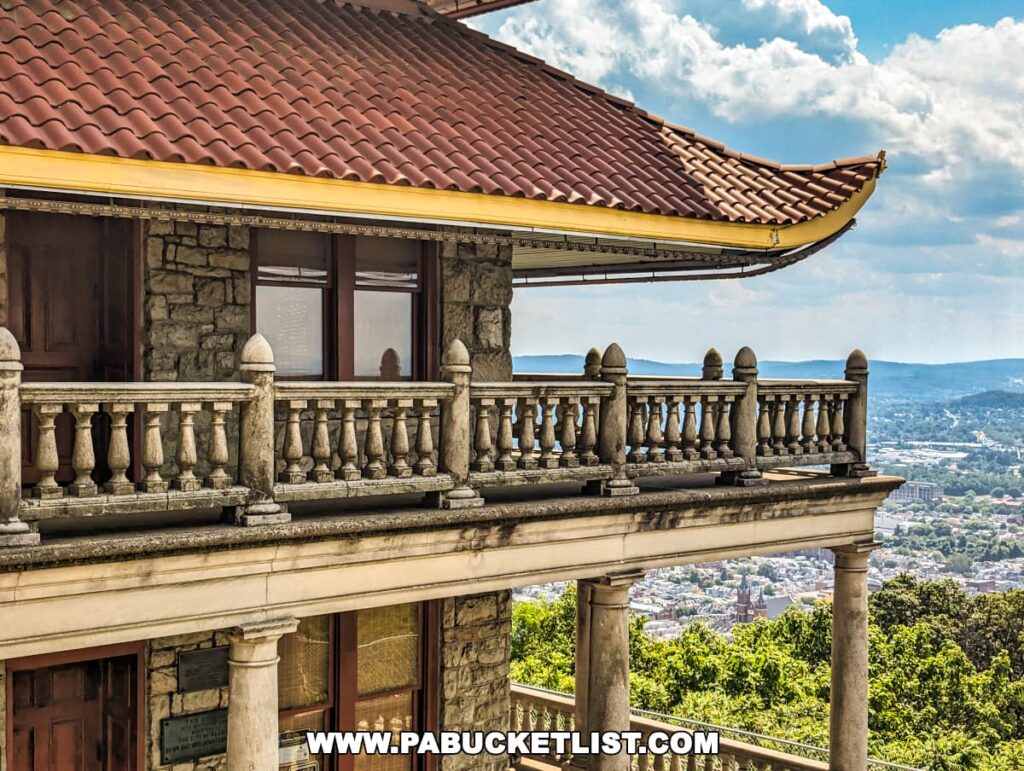 The photo provides a close-up view of the Reading Pagoda in Berks County, PA, highlighting the intricate stonework, wooden doors, and ornate balcony railings. The red tiled roof with upswept eaves is prominently featured, and the scenic backdrop includes the sprawling city of Reading and the surrounding hills under a partly cloudy sky.