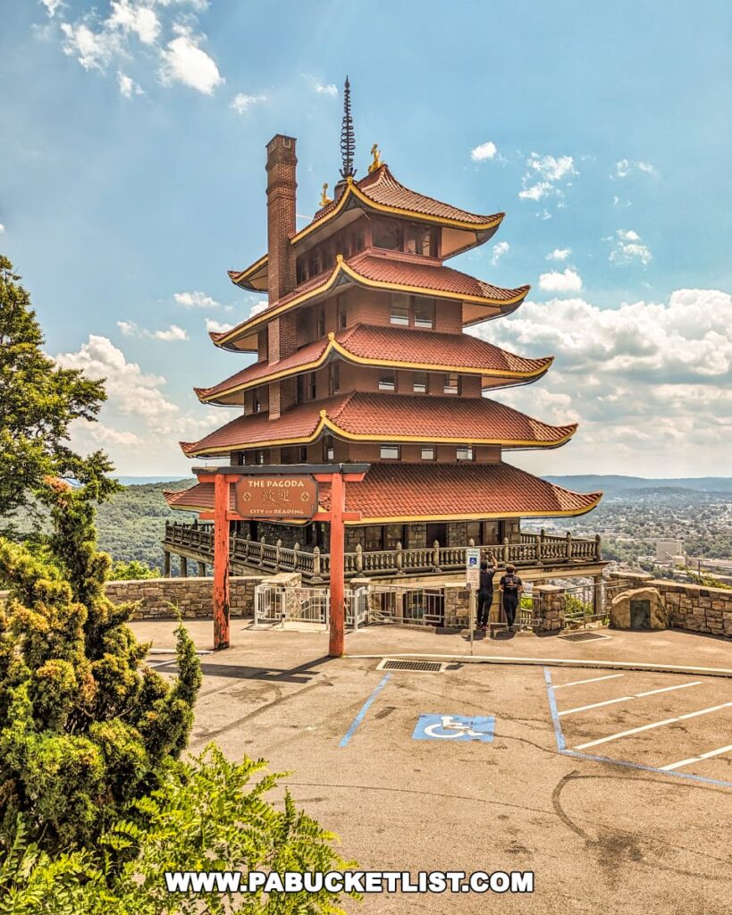 The photo features the Reading Pagoda in Berks County, PA, showcasing its seven-story structure with five distinct red roofs and upswept eaves. The torii gate at the entrance stands prominently, with a backdrop of lush greenery and the city of Reading visible in the distance under a partly cloudy sky. Two visitors are seen near the entrance, adding a sense of scale to the historic landmark.