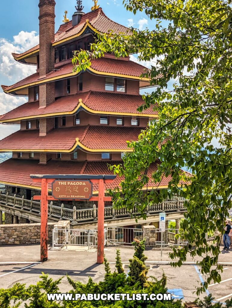 The image highlights the Reading Pagoda in Berks County, PA, with its seven-story structure and distinctive red roofs with upswept eaves. The torii gate at the entrance is prominently displayed, framed by green foliage in the foreground. The Pagoda's stone and wooden details are clearly visible, with the city of Reading seen in the background under a partly cloudy sky.