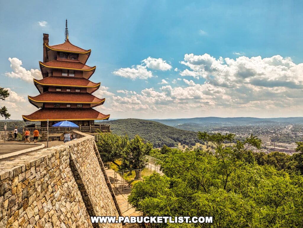 The photo shows the Reading Pagoda in Berks County, PA, perched on the southern tip of Mt. Penn. The seven-story structure with its five overhanging red roofs stands prominently, overlooking the city of Reading and the surrounding hills. Visitors can be seen near the stone wall, enjoying the scenic views on a sunny day with a clear blue sky and scattered clouds. The lush greenery adds to the serene atmosphere of the location.