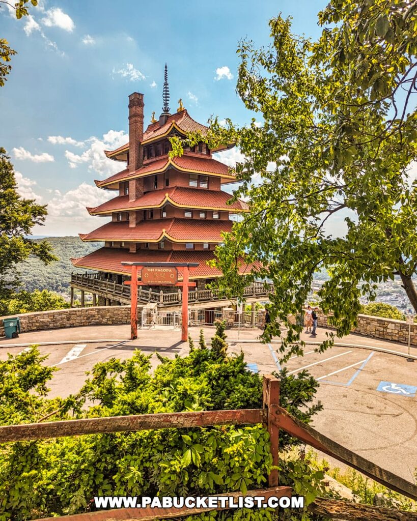 The photo depicts the Reading Pagoda in Berks County, PA, with its striking seven-story structure and five red roofs with upswept eaves. The torii gate at the entrance is visible, framed by lush green foliage in the foreground. The Pagoda stands against a backdrop of hills and a partly cloudy sky, with visitors seen near the entrance and in the surrounding area, enjoying the historic site.