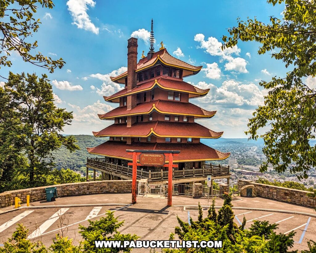 The image captures the Reading Pagoda in Berks County, PA, standing seven stories high with its distinctive five overhanging red roofs and upswept eaves. The torii gate at the entrance is prominently displayed, with lush greenery and the city of Reading visible in the background under a partly cloudy sky. The stone wall and parking area in the foreground add to the historic and serene atmosphere of the landmark.