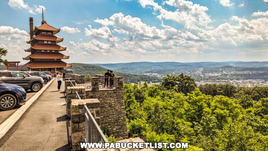 The photo features the Reading Pagoda in Berks County, PA, viewed from the parking lot. The seven-story structure with its red roofs and upswept eaves is prominently visible on the left side. Visitors are seen enjoying the scenic overlook, which offers expansive views of the lush green landscape and the city of Reading below, under a partly cloudy sky. The stone wall and parked cars add context to the accessibility and popularity of this historic site.