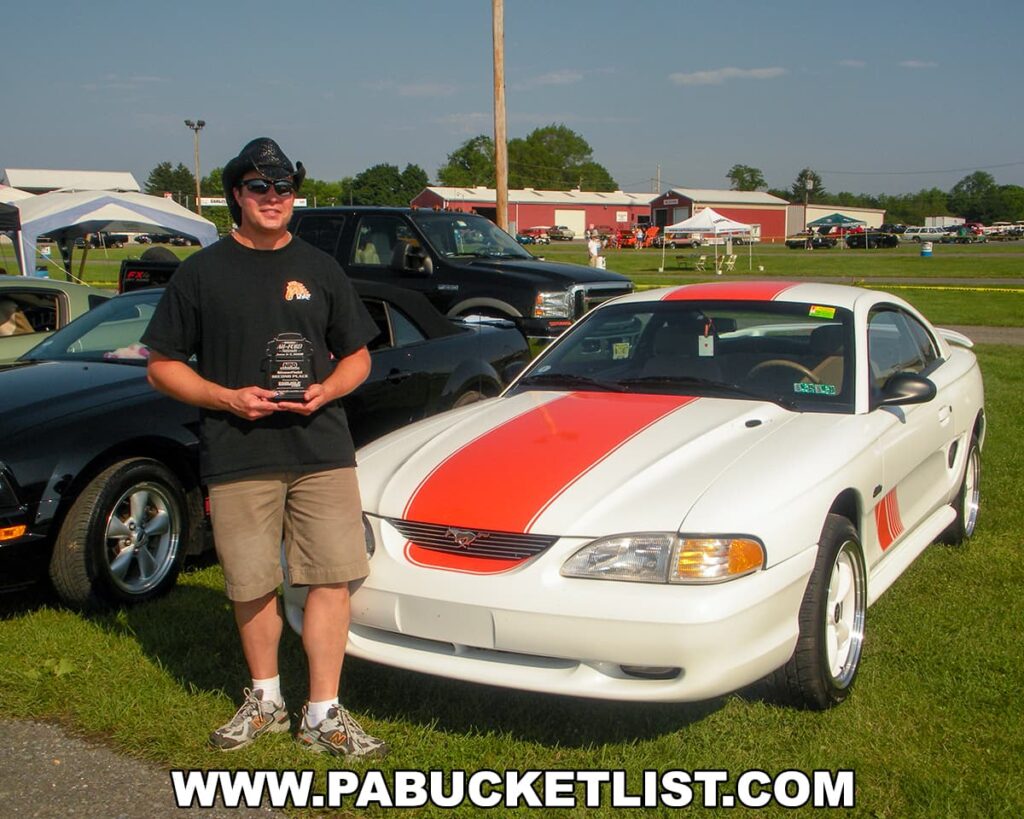 Author Rusty Glessner standing next to his white 1996 Ford Mustang GT with orange racing stripes at the Carlisle Events Ford Nationals car show in Cumberland County, PA, in 2009. He is holding an award plaque and wearing a black t-shirt, shorts, sunglasses, and a hat. Other cars and event tents are visible in the background on a sunny day, highlighting the lively atmosphere of the car show.