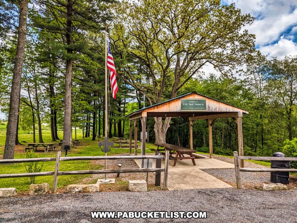 A picnic pavilion at Scripture Rocks Heritage Park in Jefferson County, Pennsylvania, located next to a parking area. The pavilion is dedicated to E.M. "Jack" Parker Jr. and features picnic tables beneath a wooden shelter. An American flag is prominently displayed nearby, and the area is surrounded by tall trees and green grass, providing a peaceful spot for visitors to rest and enjoy the park's natural beauty.