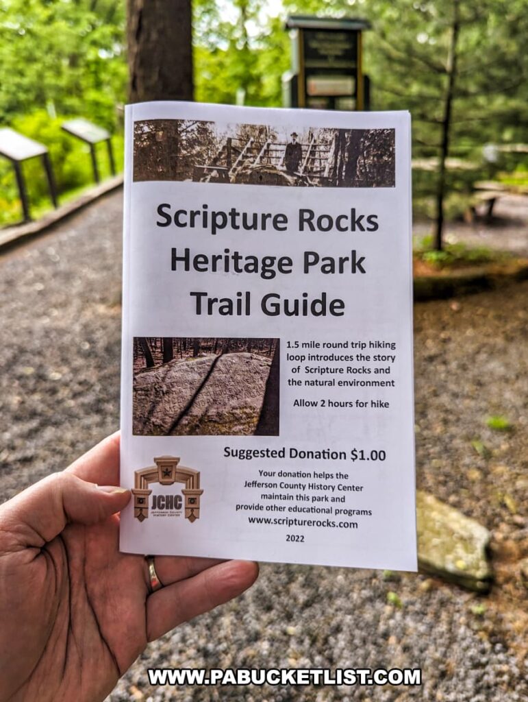 A hand holding the Scripture Rocks Heritage Park Trail Guide in Jefferson County, Pennsylvania. The guide provides information about the 1.5-mile round trip hiking loop, the history of the park, and the natural environment. The background shows an outdoor setting with educational signs and a gravel path, inviting visitors to explore the park and its historical features. The guide suggests a donation to help maintain the park and support educational programs provided by the Jefferson County History Center.
