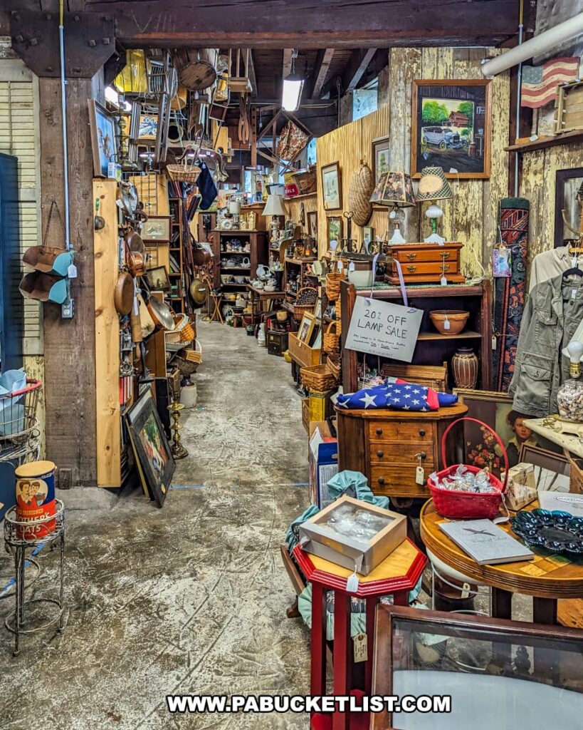A narrow aisle in the basement level of the Strasburg Antique Market in Lancaster County, filled with a diverse array of vintage and collectible items, including framed art, kitchenware, furniture, and decorative pieces. The space is warmly lit and features wooden beams and rustic walls, enhancing its charming, nostalgic atmosphere.