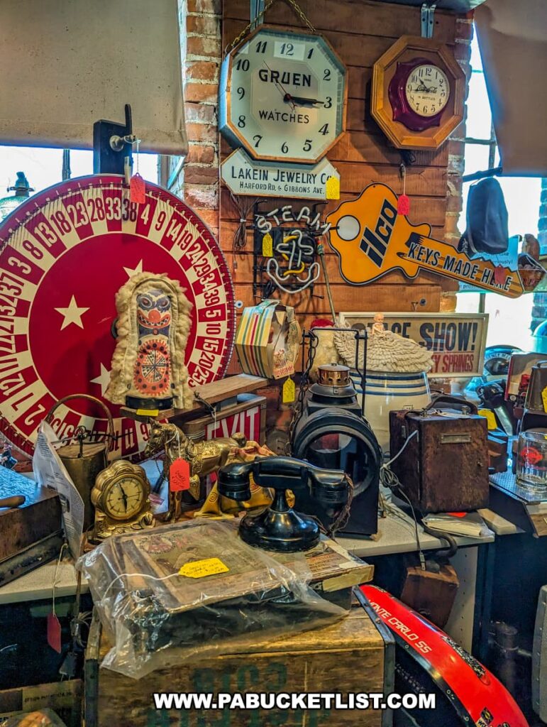 A vibrant display at the Strasburg Antique Market featuring a variety of vintage items, including a large red and white roulette wheel, antique clocks, a retro telephone, old signage, and various collectibles. The eclectic assortment is arranged on a table and against a brick wall, showcasing the market's diverse offerings and historical charm.