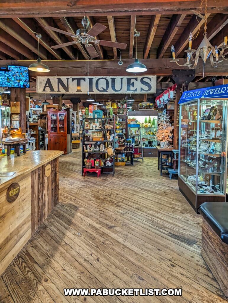 The interior of the Strasburg Antique Market, featuring rustic wooden floors and exposed beams that enhance its historical charm. The market is filled with neatly organized displays of antiques, including furniture, glassware, estate jewelry, and various collectibles. A large "Antiques" sign hangs prominently, guiding visitors through the diverse selection. Warm lighting and ceiling fans contribute to the cozy and inviting atmosphere, making it an enjoyable space to explore and discover unique treasures.