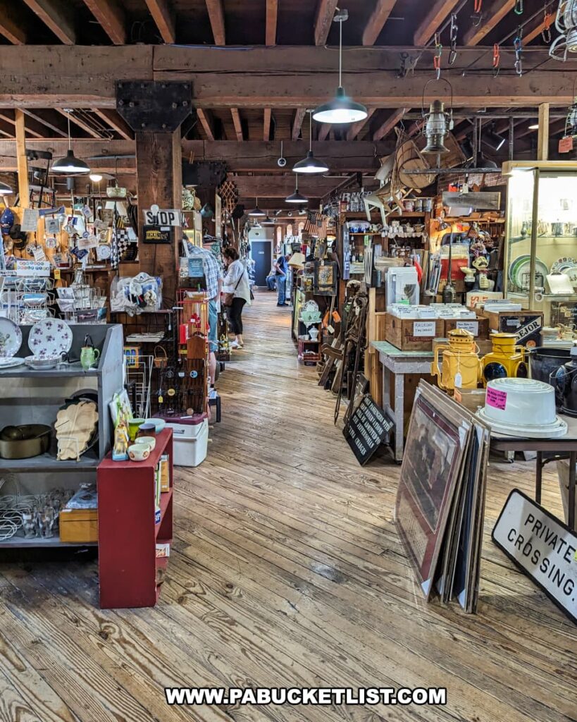 A bustling aisle inside the Strasburg Antique Market, showcasing a variety of antiques and collectibles displayed on both sides. The rustic wooden floors and exposed beams add to the historical charm of the restored 1898 tobacco warehouse. Shoppers are seen browsing through the diverse selection, which includes dishes, vintage signs, and various knick-knacks. Warm lighting from hanging lamps creates a cozy and inviting atmosphere, enhancing the overall shopping experience.