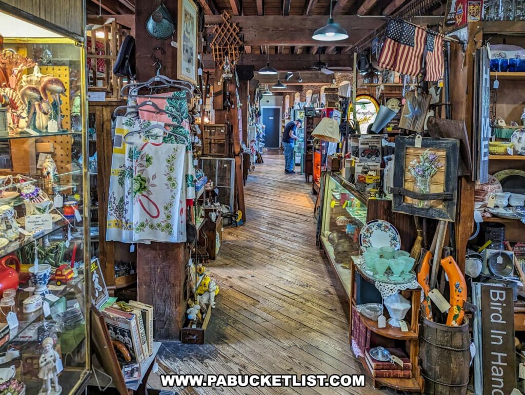 A vibrant and busy aisle at the Strasburg Antique Market, showcasing a diverse range of antiques and collectibles. Items are neatly arranged on shelves and hangers, including vintage clothing, decorative plates, glassware, books, and patriotic memorabilia. The rustic wooden floors and exposed beams enhance the historical atmosphere of the restored 1898 tobacco warehouse. Shoppers are seen exploring the various displays, which are illuminated by warm hanging lights, creating a welcoming and nostalgic environment.