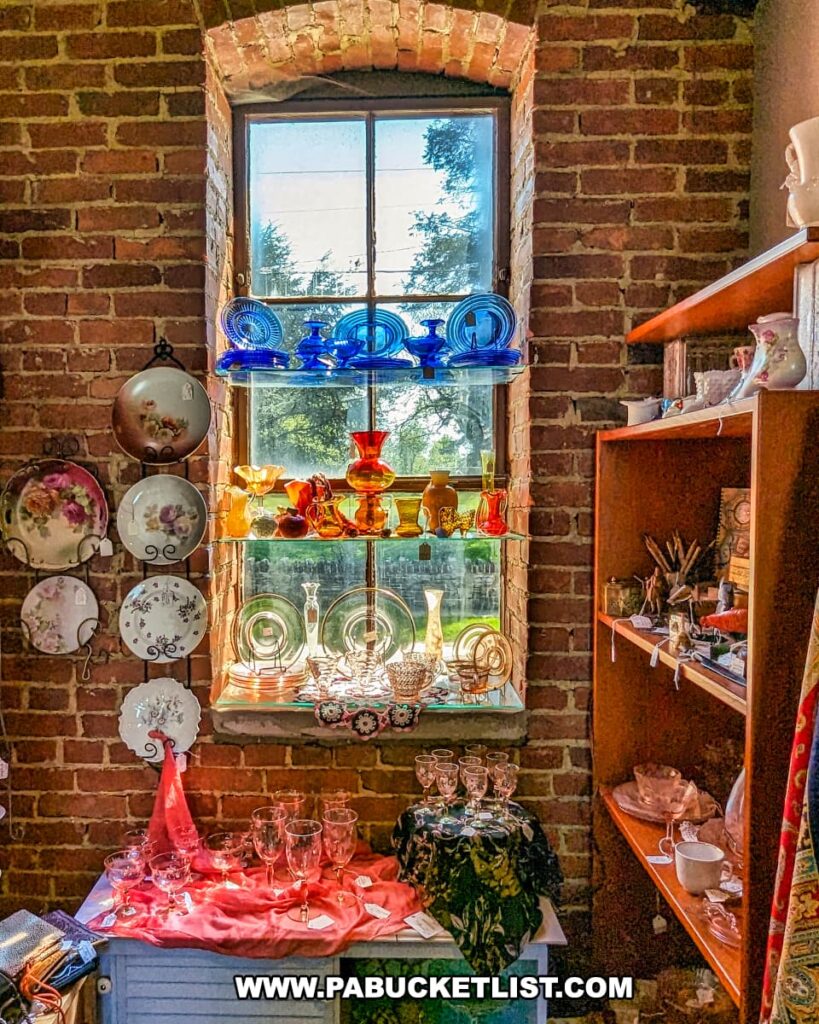 A charming display of vintage glassware at the Strasburg Antique Market, set against a brick wall with a large window. The sunlight illuminates the colorful glass pieces, including blue, red, and amber items, creating a vibrant and inviting scene. Surrounding the window are decorative plates with floral designs, and additional glassware is arranged on a table and shelves nearby. The rustic brick and natural light enhance the nostalgic and historical ambiance of the market.