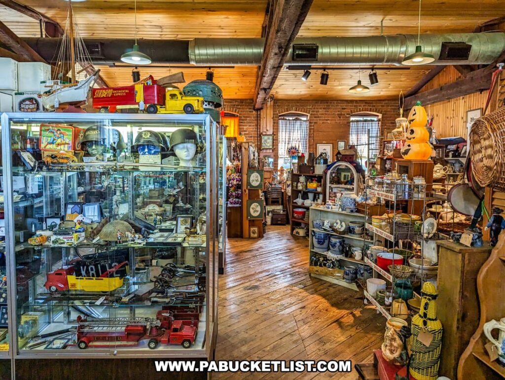 A diverse display area at the Strasburg Antique Market featuring vintage military memorabilia and other collectibles. The glass case on the left showcases items such as helmets, model ships, and toy trucks, while shelves and tables around the room are filled with various antiques, including pottery, glassware, and holiday decorations. The room's rustic charm is enhanced by wooden floors, exposed beams, and natural light from the windows. The eclectic mix of items and the inviting setup encourage visitors to explore and discover unique treasures.
