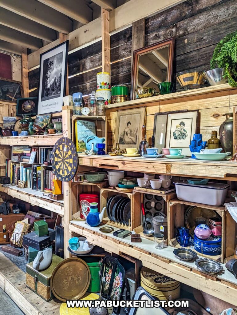 A display at the Tollbooth Antique Warehouse in Lancaster County, Pennsylvania, features wooden shelves filled with a variety of vintage and collectible items. The shelves hold an assortment of books, framed artwork, decorative glassware, teapots, and retro kitchenware. Other items include a dartboard, old tins, and various knick-knacks, showcasing the diverse range of antiques and memorabilia available at the multi-vendor store. The rustic wooden walls and exposed beams add to the charm and character of the former factory building, creating an inviting atmosphere for treasure hunters.