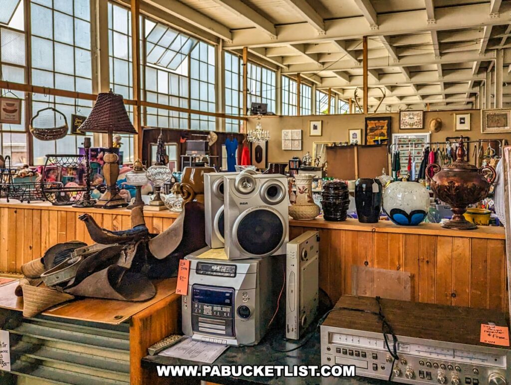 The interior of the Tollbooth Antique Warehouse in Lancaster County, Pennsylvania, features a display of vintage electronics and eclectic decor. Prominently placed is an RCA stereo system with large speakers, surrounded by various antique items including lamps, vases, and decorative pottery. The background showcases an array of additional vintage and collectible items, such as woven baskets, framed artwork, and clothing racks. The spacious area is lit by natural light from large industrial windows, highlighting the expansive and diverse collection available at the multi-vendor antique store located in a former factory building.