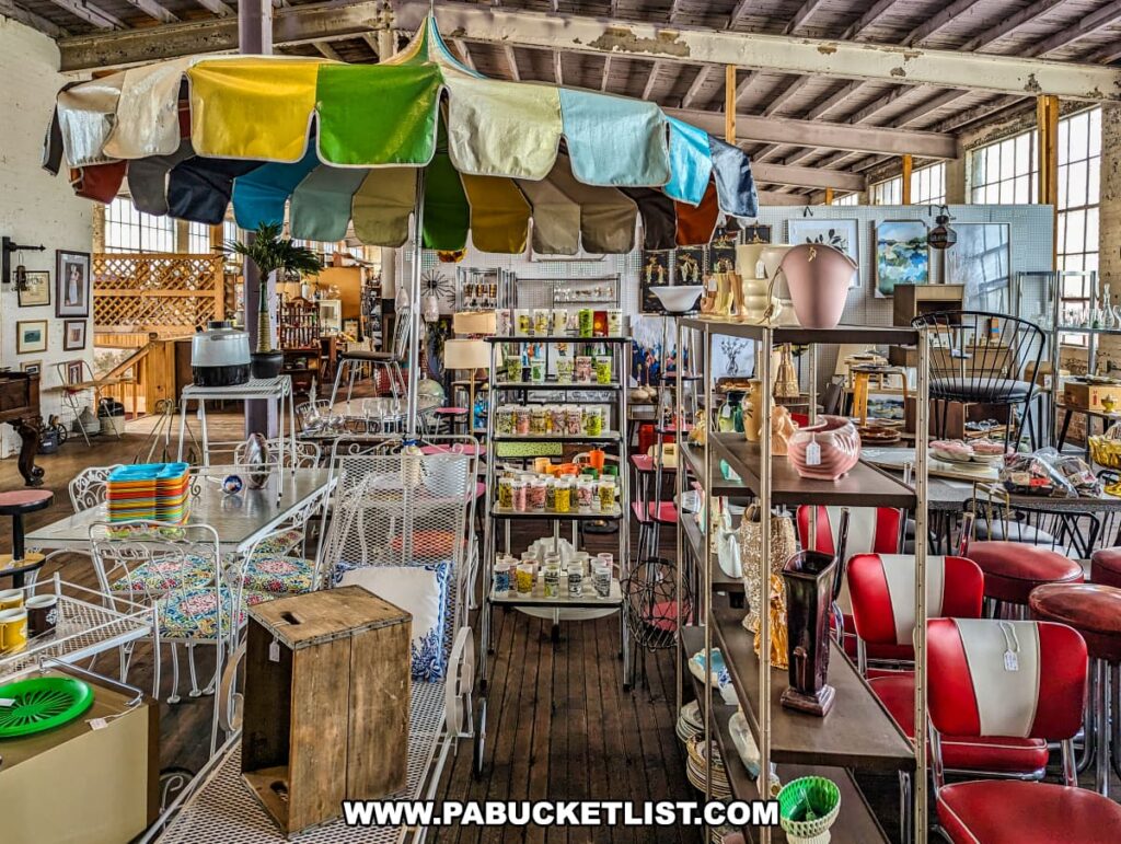 A vibrant section of the Tollbooth Antique Warehouse in Lancaster County, Pennsylvania, displays a colorful assortment of vintage lawn furniture and other collectibles. The scene includes a metal patio set with white chairs and a table, topped with a multi-colored patio umbrella. Surrounding the furniture are shelves filled with various items such as vases, lamps, kitchenware, and decor pieces. The area is well-lit by large industrial windows, highlighting the eclectic mix of vintage and retro items available at this expansive multi-vendor antique store located in a former factory building.