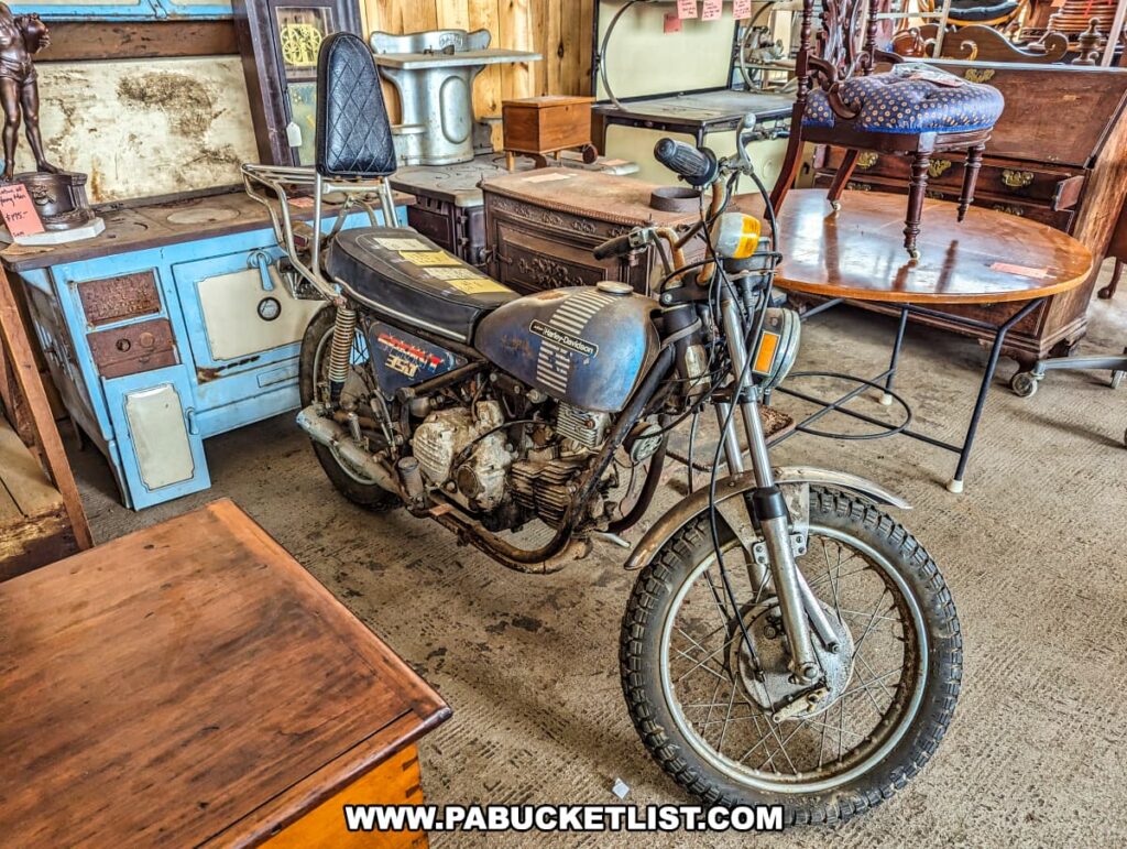 Inside the Tollbooth Antique Warehouse in Lancaster County, Pennsylvania, a vintage motorcycle is prominently displayed among an assortment of antique furniture. The motorcycle, with its weathered blue paint and retro design, is positioned next to various wooden tables, a blue antique stove, and other vintage pieces. The eclectic mix of items highlights the diverse offerings at this expansive multi-vendor antique store located in a former factory building. The setting provides a nostalgic glimpse into the past, with unique treasures waiting to be discovered.