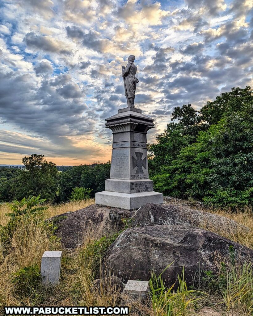 The photo shows the monument to the 155th Pennsylvania Volunteers on Little Round Top in Gettysburg National Military Park. The statue depicts a Union soldier standing at ease with a rifle, atop a stone pedestal inscribed with details of the regiment. The monument is set on a rocky outcrop surrounded by grass and trees, with a dramatic sky filled with scattered clouds illuminated by the rising sun in the background. The scene captures the tranquil and reflective atmosphere of the historic battlefield.