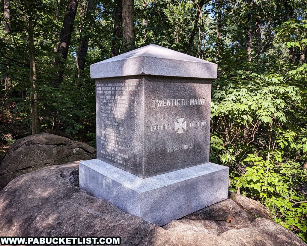 The photo shows the monument dedicated to the 20th Maine Volunteer Infantry Regiment at Gettysburg National Military Park. The monument is a simple, rectangular stone structure with inscriptions honoring the regiment's contributions and sacrifices. It is situated on a large rock surrounded by a wooded area, with lush green foliage and trees providing a serene and reflective backdrop. The setting highlights the historical significance and natural beauty of this iconic location on the battlefield.