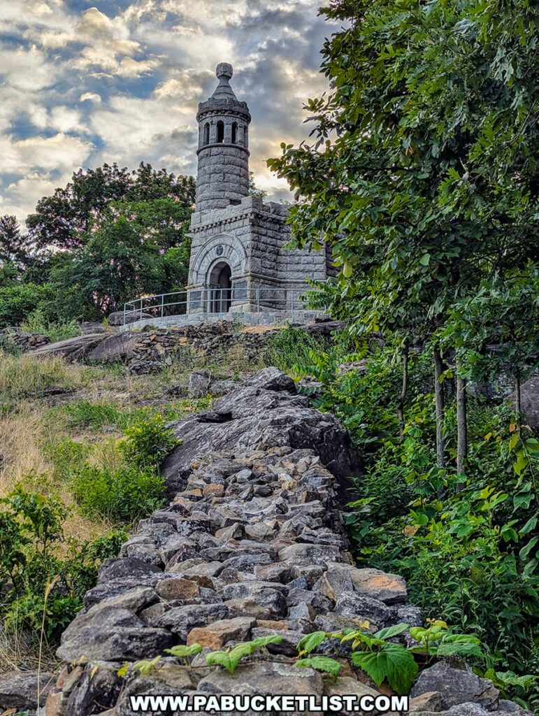 The photo shows the 44th New York Infantry Monument, a small castle-like structure, situated on Little Round Top in Gettysburg National Military Park. The monument is made of stone and features a rounded turret with an arched entrance. The structure is perched atop a rocky hill, with a rugged stone path leading up to it. The surrounding area is lush with greenery, and the sky above is partly cloudy, capturing the serene yet solemn atmosphere of the historic battlefield at sunrise.