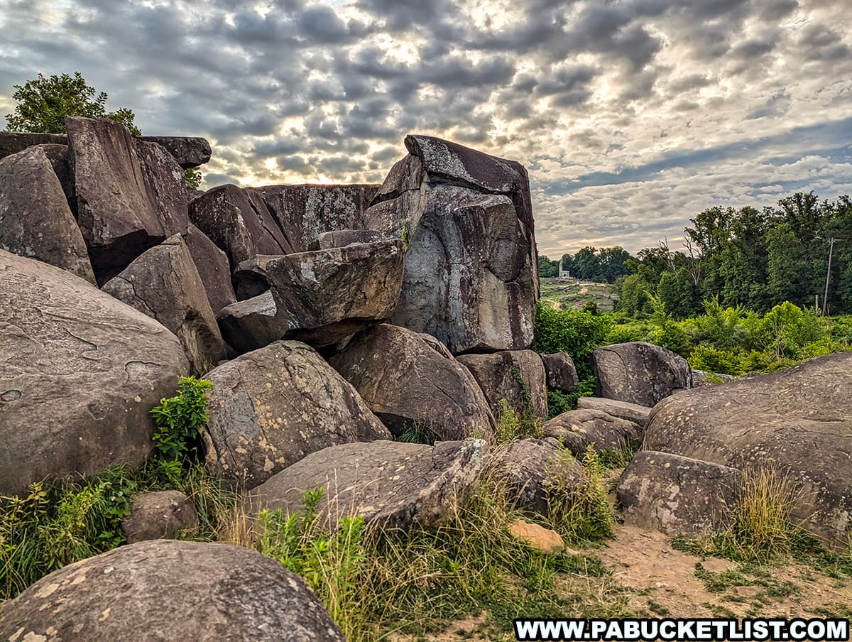 The photo shows the rocky terrain of Devil's Den in Gettysburg National Military Park. Large boulders are piled together, creating a rugged and imposing landscape. The sky is partly cloudy, with sunlight filtering through, casting shadows on the rocks. Surrounding the boulders is a mix of green foliage and grass, adding a natural contrast to the stone. The scene evokes the historical significance and natural beauty of this notable battlefield location.