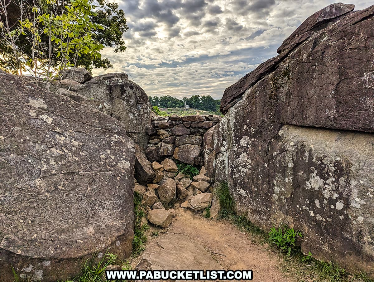The photo shows the sniper's nest at Devil's Den in Gettysburg National Military Park. Large, weathered boulders form a natural enclosure, with a small stone wall built between them. The ground is dirt and scattered with smaller rocks, and a few green plants grow in the crevices. The sky is partly cloudy, allowing sunlight to filter through and illuminate the scene. This historical location is both somber and captivating, highlighting the rugged terrain where significant events took place during the Battle of Gettysburg.
