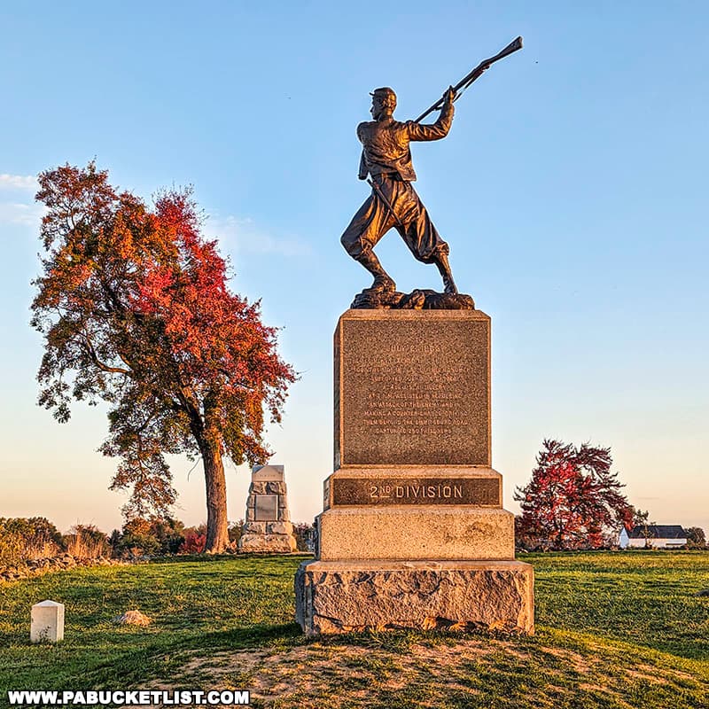 The photo features a monument on Cemetery Ridge in Gettysburg National Military Park, depicting a Union soldier in a dynamic pose, readying his rifle. The statue stands on a tall stone pedestal inscribed with details of the 2nd Division. The scene is set against a clear blue sky with vibrant autumn foliage, including a tree with red and orange leaves. Additional monuments can be seen in the background, along with a glimpse of the battlefield's expansive grassy area. The image captures the historical significance and natural beauty of the site.