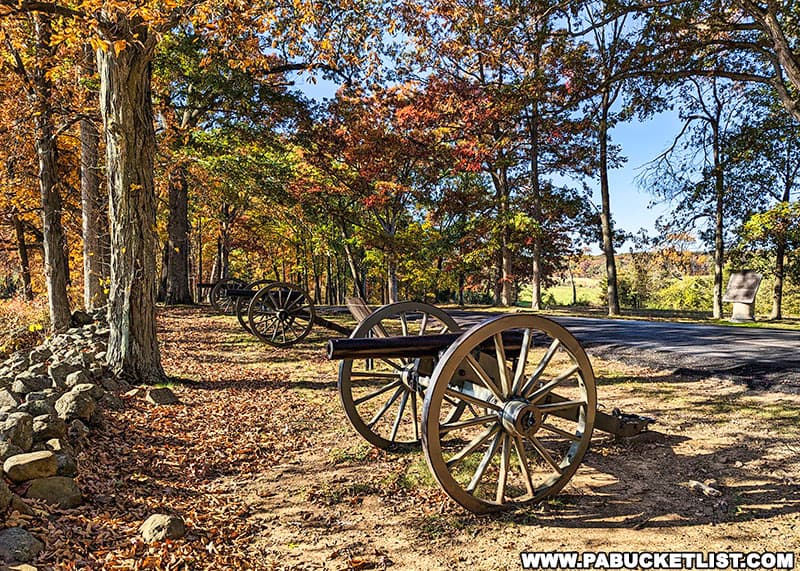 The photo shows a row of historic cannons along Confederate Avenue in Gettysburg National Military Park. The scene is set during autumn, with trees displaying a vibrant mix of orange, red, and yellow foliage. Fallen leaves cover the ground around the cannons and a stone wall runs parallel to the line of cannons. The clear blue sky and the dappled sunlight filtering through the trees create a picturesque and serene atmosphere, highlighting the historic significance and natural beauty of the battlefield.