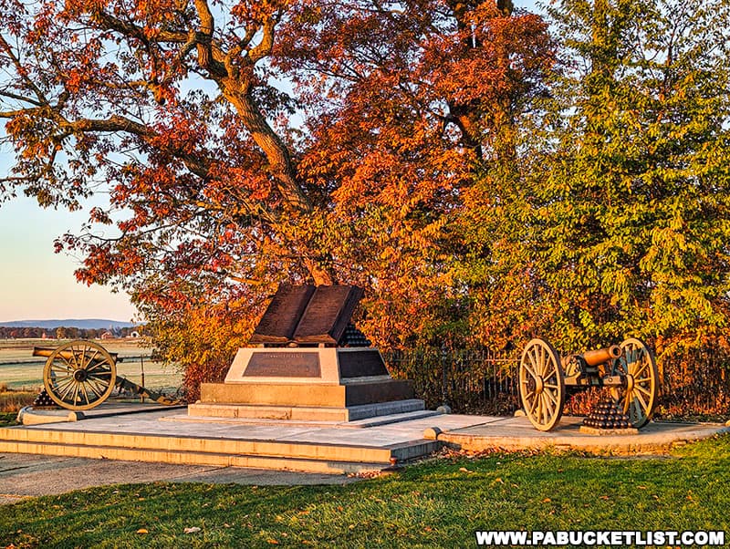 The photo captures the High Water Mark of the Confederacy monument at Gettysburg National Military Park, surrounded by vibrant autumn foliage. The monument consists of two large plaques mounted on a stone base, flanked by historic cannons on either side. The trees in the background display brilliant red and orange leaves, creating a striking contrast with the monument's stone and metal elements. The scene is set against a backdrop of open fields and distant hills under a clear sky, highlighting the historical importance and natural beauty of this significant location.