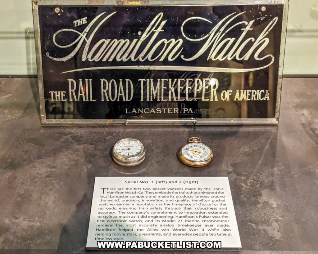An exhibit at the National Watch and Clock Museum in Lancaster County, Pennsylvania, featuring two early pocket watches made by the Hamilton Watch Company. The display includes a vintage sign that reads "The Hamilton Watch, The Rail Road Timekeeper of America, Lancaster, PA." Below the sign, the pocket watches are placed side by side. The left watch is identified as Serial No. 1, and the right watch as Serial No. 2. An informational plaque details the historical significance of these watches, highlighting Hamilton's reputation for precision, innovation, and quality. The plaque also mentions Hamilton's contribution to railroad safety and its role in supporting the Allies during World War II. The exhibit emphasizes the craftsmanship and technological advancements of Hamilton watches.