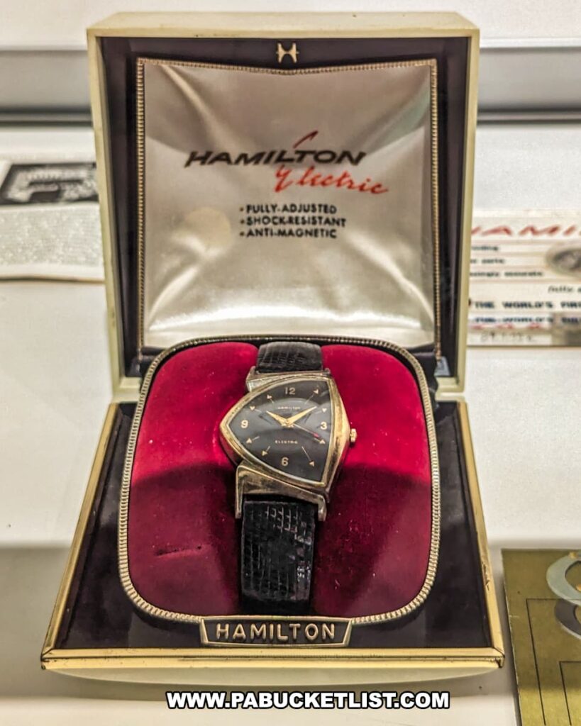 A display at the National Watch and Clock Museum in Lancaster County, Pennsylvania, featuring a vintage Hamilton Electric watch. The watch, presented in an elegant case with a red velvet lining, has a distinctive triangular black face with gold accents and a black leather strap. The case lid is lined with satin and bears the Hamilton logo along with the words "Hamilton Electric," highlighting the watch's attributes such as being fully adjusted, shock-resistant, and anti-magnetic. The background includes informational materials about the watch, showcasing its historical significance and innovative design.