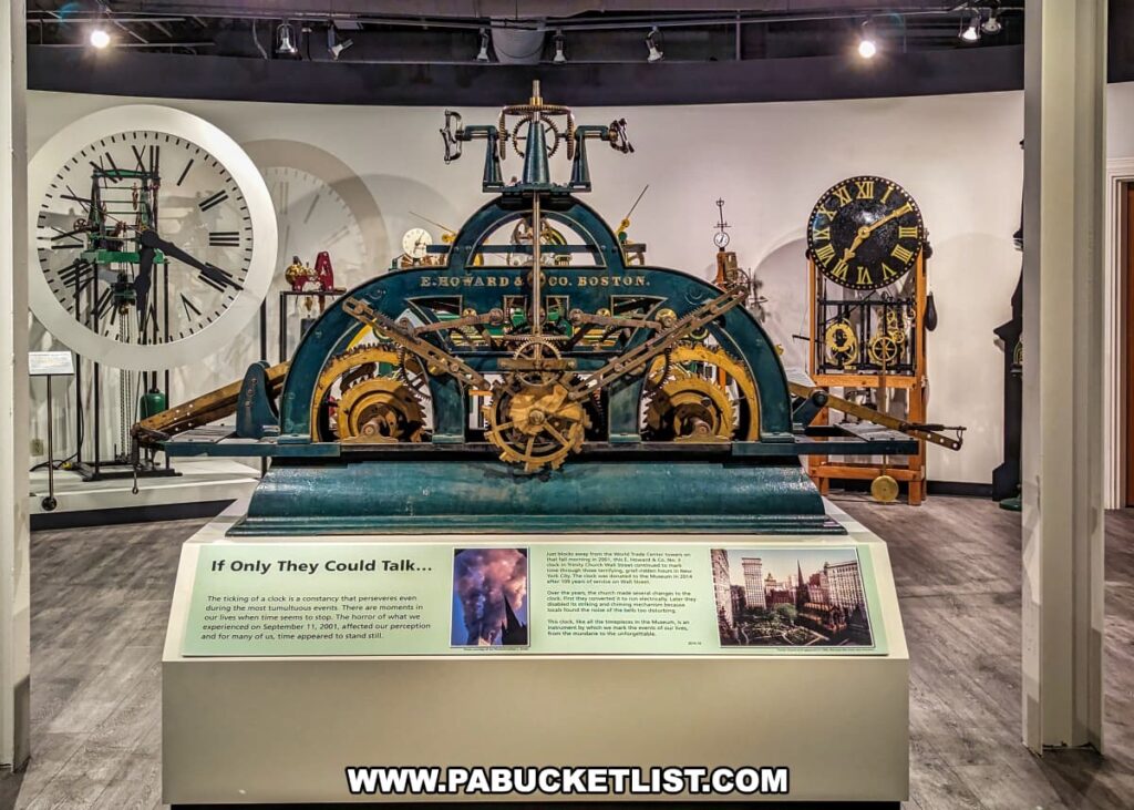 A display at the National Watch and Clock Museum in Lancaster County, Pennsylvania, showcasing a large, early clock mechanism made by E. Howard & Co. of Boston. The intricate machinery features a complex arrangement of gears, levers, and pulleys, all housed in a robust metal frame. Behind the clock mechanism, several large clock faces and additional timekeeping devices are visible, demonstrating the evolution of clock technology. An informational plaque in the foreground provides context about the significance of the clock, noting its historical importance and connection to notable events, including the impact of time perception during September 11, 2001. The exhibit is well-lit, highlighting the detailed engineering and craftsmanship of the clock.
