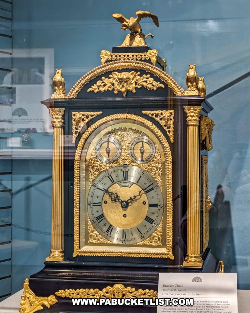 An ornate bracket clock on display at the National Watch and Clock Museum in Lancaster County, Pennsylvania. The clock, made by Thomas R. Russell from Liverpool, England around 1880, features intricate gilded decorations. The clock face has Roman numerals and two smaller dials above it. The casing is black with gold detailing, including columns on either side, floral patterns, and a majestic eagle perched on top. Two small owl figurines sit on either side of the top. A descriptive plaque provides information about the clockmaker and the history of the timepiece. The clock is showcased in a glass display case with a blue background.