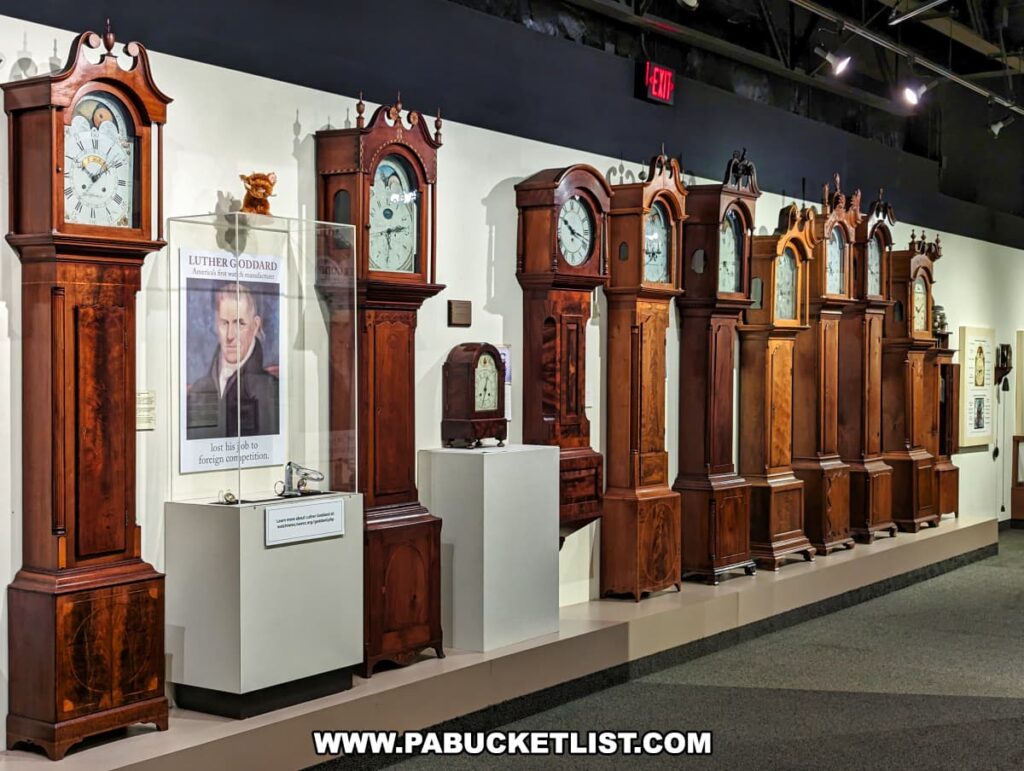 A display of numerous grandfather clocks at the National Watch and Clock Museum in Lancaster County, Pennsylvania. The clocks, with their tall wooden cases and ornate designs, are lined up against a white wall. Each clock features a distinct face with Roman numerals and various decorative elements. A poster of Luther Goddard, an American clockmaker, is displayed in a glass case along with some small artifacts. The exhibit is well-lit, allowing visitors to appreciate the fine craftsmanship and historical significance of each grandfather clock. The arrangement highlights the evolution of design and technology in clockmaking.