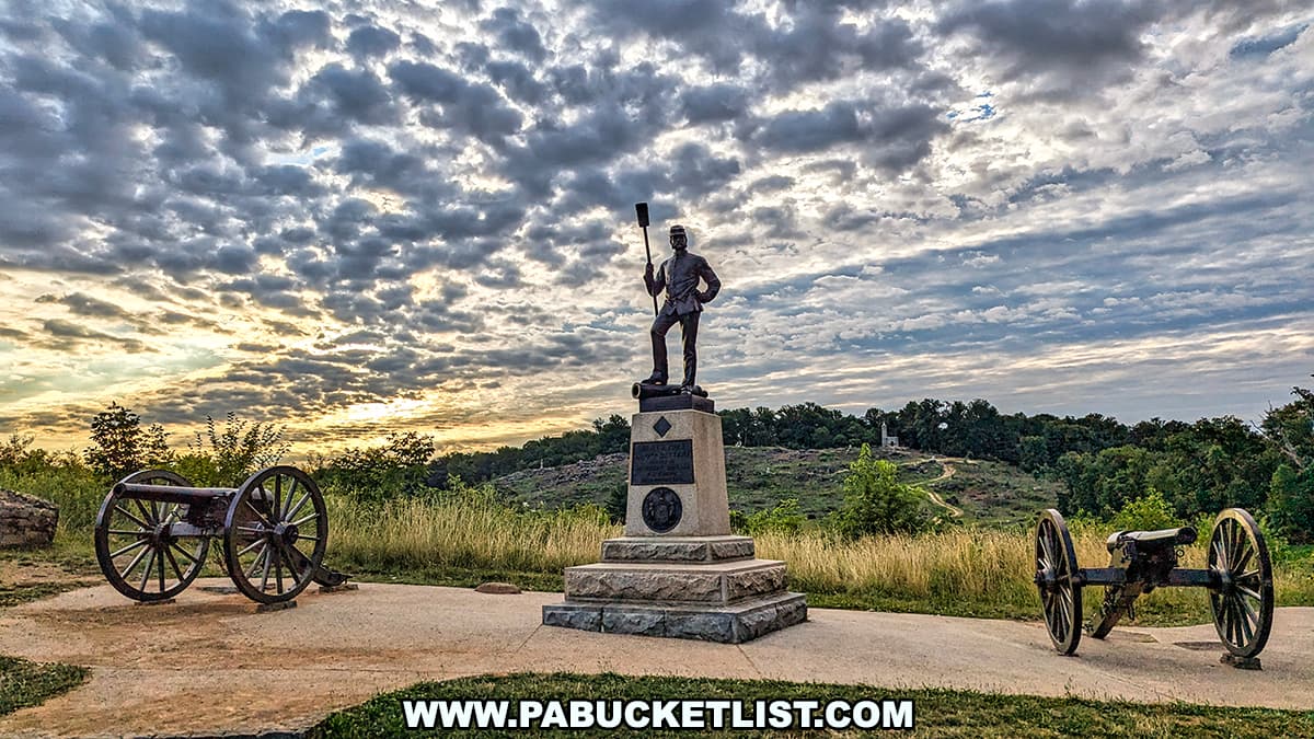 The photo shows a monument and cannons near Devil's Den in Gettysburg National Military Park. The monument features a statue of an artilleryman holding a cannon rammer, standing on a stone pedestal inscribed with commemorative details. The scene is framed by two cannons on either side of the monument. The background includes a lush green landscape with trees and rocky terrain under a dramatic, cloud-filled sky illuminated by the early morning light. The setting evokes a sense of historical significance and serene beauty at this notable battlefield location.