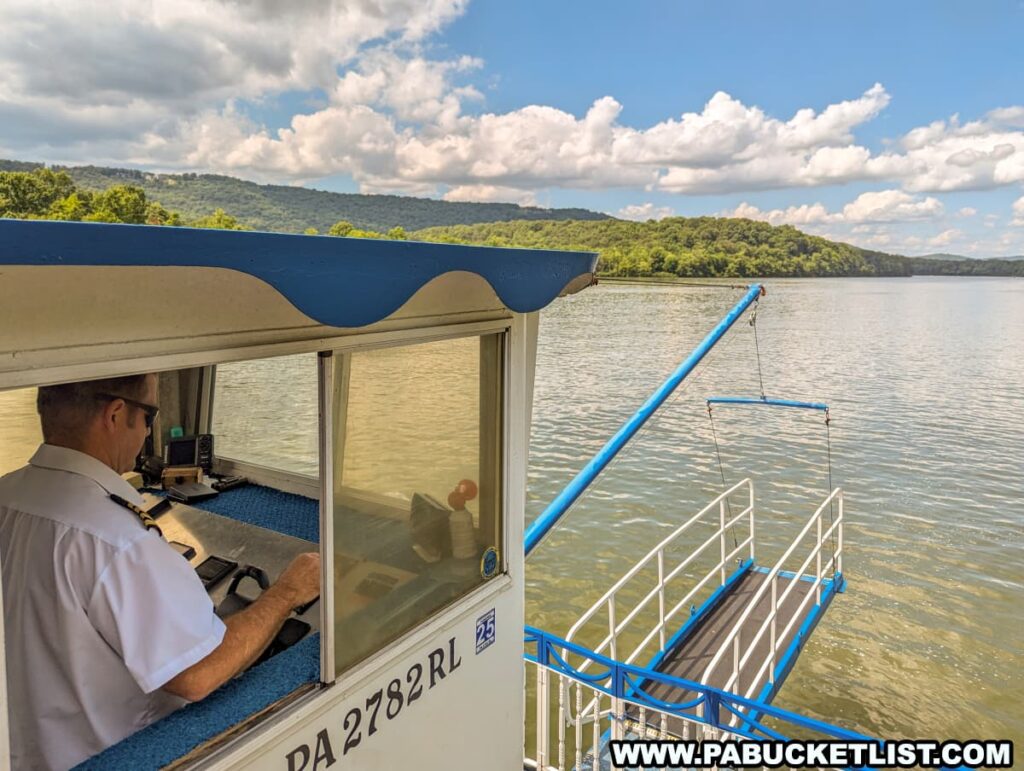 A photo of the captain steering the Proud Mary Showboat on Raystown Lake. The image shows the captain in the control room, with large windows offering a view of the calm lake and surrounding hills under a partly cloudy sky. The boat's boarding ramp is visible extending over the water.