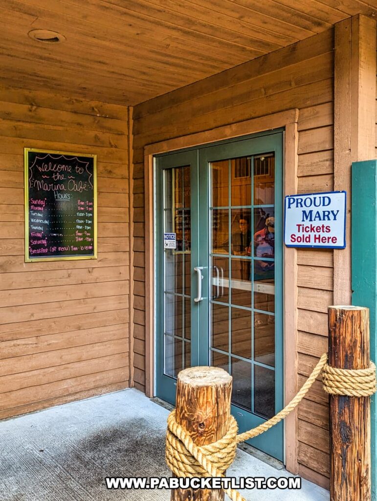 The entrance to the Marina Café at Raystown Lake, featuring double glass doors and a wooden exterior. A sign next to the door indicates that Proud Mary tickets are sold here. A chalkboard on the wall lists the café's hours, and decorative wooden posts with rope add a nautical touch to the entrance.