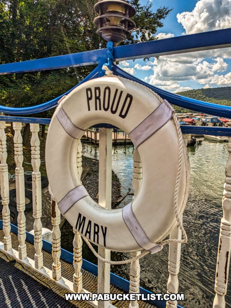 A life preserver labeled "Proud Mary" is mounted on the railing of the showboat, with the scenic Raystown Lake and surrounding green hills visible in the background under a partly cloudy sky.