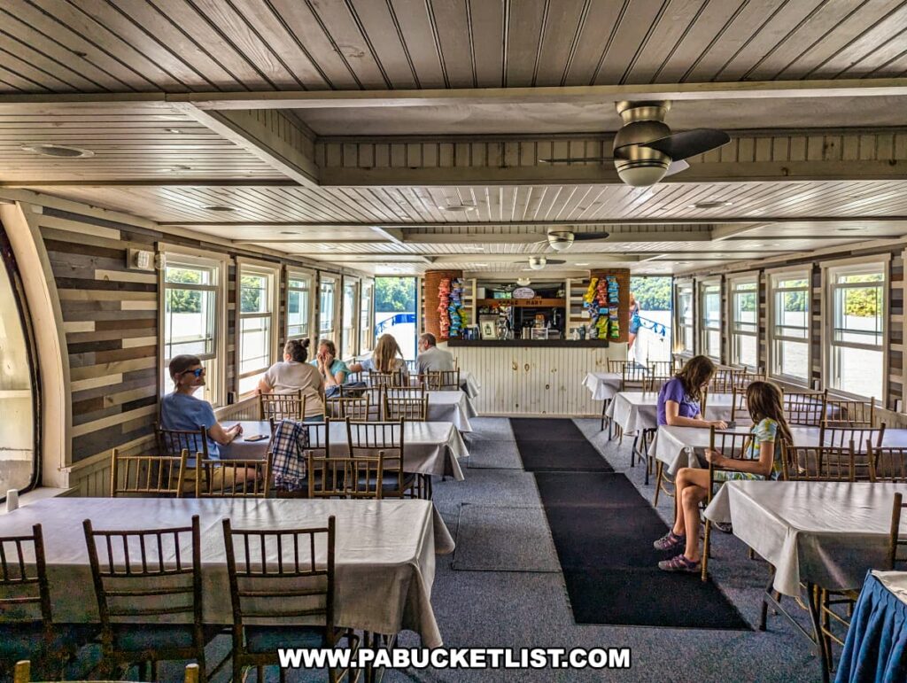Passengers seated in the lower level dining area of the Proud Mary Showboat, with tables covered in white tablecloths and wooden chairs. The area is brightened by large windows offering views of Raystown Lake. The snack bar is visible at the far end of the room, adorned with colorful chip bags. The ceiling and walls feature a wooden finish, creating a cozy atmosphere.