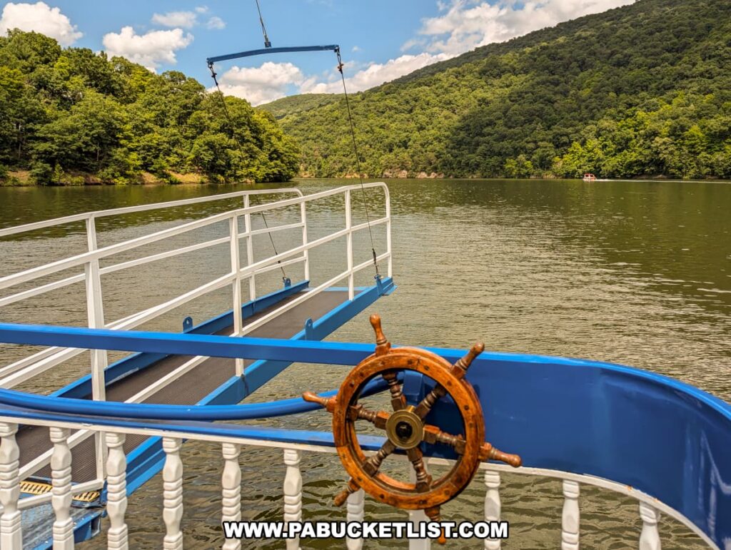 A view from the Proud Mary Showboat showing the boarding ramp extending over Raystown Lake. The scene includes a decorative wooden ship's wheel mounted on the railing, with calm water and green, forested hills in the background under a partly cloudy sky.